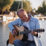 Mies istuu ja hymyilee sekä soittaa kitaraa taustalla merimaisema.
